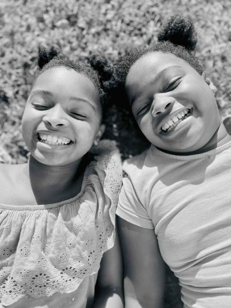 A black and white image of two smiling young girls laying on the grass with their bodies close together. Photo by Fikelphi Ndsisile via Pexels.com